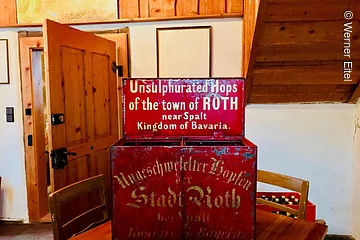 Eine rote Kiste aus Metall steht auf eibnem Tisch in einem Wohnraum. Auf der Kiste steht in weißer Schrift: Unsulphurated Hops of the town of Roth near Spalt Kingdom of Bavaria