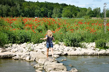 Ein junges Mädchen läuft über eine Steinreihe die durch einen Fluss führt. Dahinter ein rot blühendes Mohnblumenfeld.