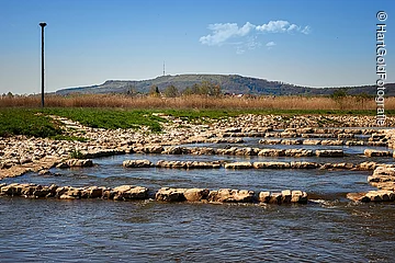 Ein breiter flacher Bach fließt durch eine grüne Landschaft. Im Bach eine Fischtreppe aus Steinen. Am Ufer des Baches liegen Steine.