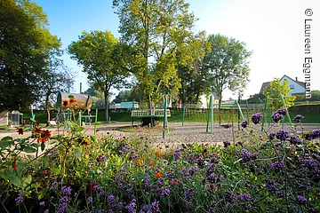 Ein Spielplatz mit Klettergerüst und Nestschaukel. Im Hintergund grüne Bäume. Im Vordergund eine bunte Blumenwiese.