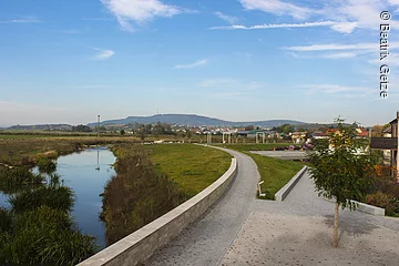Fußgängerweg am Randes eines Bachs. Am Ufer grüne Wiesen.