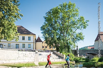 Eine Frau und ein Mann gehen über die Sulz hierfür sind einzelne Steine fußbreit im Wasser. Im Hintergrund sieht man zwei Gebäude, einen grünen Baum der den Beginn einer steinernen Brücke rechts markiert.
