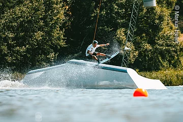 Ein Wakeboarder fährt über ein Hindernis im Wasser. Dahinter am Ufer grüne Bäume und Sträucher.