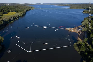 Luftbild von der Wakeboardanlage. Im See Hindernisse für die Wakeboardfahrer.  Am See ein Strand mit vielen Besuchern. Der See ist umgeben von grünen Bäumen und Sträuchern.