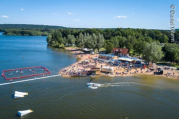 Luftbild von der Wakeboardanlage. Im See Hindernisse für die Wakeboardfahrer.  Am See ein Strand mit vielen Besuchern. Dahinter grüne Bäume und Sträucher.