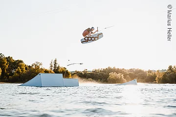 Ein Mann mit Wakeboard springt über ein Hinderniss im Wasser. Dahinter am Uder Bäume und Sträucher.