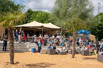 Eine Tribüne aus Stein dirket an einem Sandstrand. Darauf sitzen viele Menschen. Dahinter ein Sonnensegel und grüne Bäume.