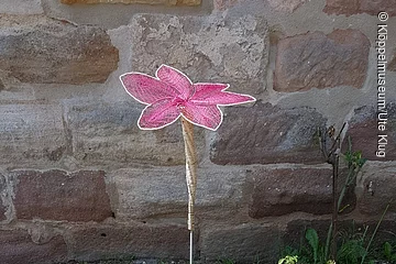 Eine geklöppelte rosa Blume steht in einer Grünfläche vor einer Mauer.