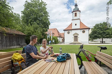 Ein Mann und eine Frau machen vor einer Kirche Pause an einem Holztisch. Auf dem Tisch liegen Helme, ein Fahrrad steht daneben.