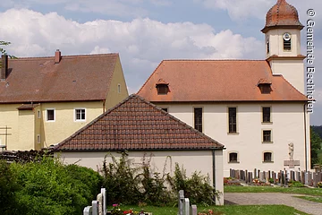 Eine Kirche mit Friedhof. Eine Kirche mit heller Fassade und kelinem Türmchen. Davor ein Friedhof mit Grabsteinen.