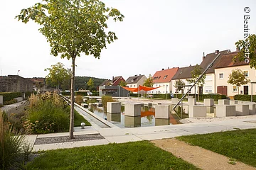 Ein Park mit einem Teich. Im Teich sind kreisförmig Steinblöcke angeordnet. Sie bidlen eine Sonnenuhr. Um den Teich Sträucher und Blumen. Im Hintergund Wohnhäuser.