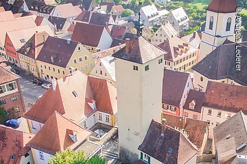 Luftbild eines Stadtturms. An den Turm angebaut ein kleines Häuschen mit grünen Fensterläden. Der Turm steht auf einer grünen Wiese umrandet von einer Mauer. Der Turm steht in einer Altstadt mit vielen Häusern.