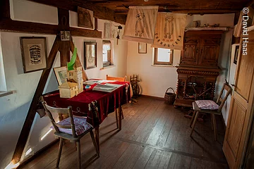 Blick in eine alte bürgerliche Stube. Fachwerkwand mit weißen Wänden. Holzboden und Holzmöbel. In der rechten Ecke ein alter Holzschrank. Links ein Tisch mit zwei Stühlen.