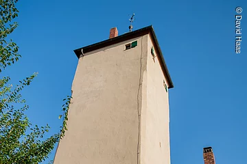 Blick von unten auf einen hohen Turm mit beiger Fassade. Daneben ein Abschnitt eines Hauses mit gleicher Fassade, einem Fenster und grünem Fensterladen.