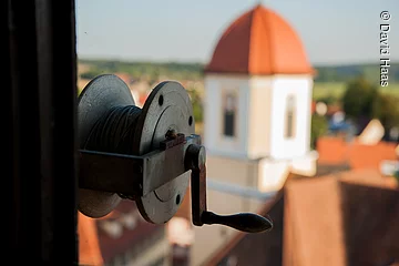 Im Fokus eine Spule mit Seil und Kurbel. Im Hintergund ist verschwommen ein Turm zu erkennen. Der Turm hat eine helle gelbe Fassade und ein rotes Haubendach.