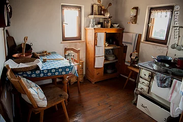 Eine kleine altertümliche Küche. Sitzecke mit zwei Stühlen in der rechten Ecke. in der hinteren Ecke ein Holzschrank. Rechts davon ein alter Holzofen zum Kochen.