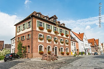 Ein großes Haus mit vier Ebenen. Ein Haus mit Steinfassade. An den Fenstern grüne Fensterläden. Daran Blumenkästen mit roten Blumen. Das Haus hat ein Walmdach. Das Haus liegt an einer gepflasterten Straße.