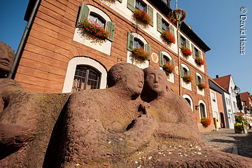 Blick und untern auf zwei Figuren eines Steinbrunnen. Die zwei Figuren halten Hände. Im Hintergund ein großes Haus mit Steinfassade. An den Fenstern grüne Fensterläden und Blumenkästen mit roten Blumen.