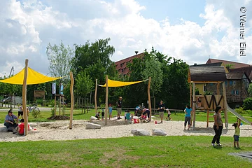 Ein Spielplatz auf Schotterfläche umgeben mit grüner Wiese. Zwei kleine gelbe Sonnensegel. Rechts daneben eine Rutsche. Kinder und Eltern spielen auf dem Spielplatz. Im Hintergund Büsche und Bäume.