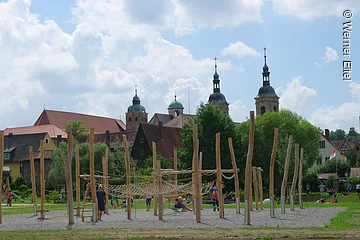 Im Vordergrund ein Spielplatz auf einer Schotterfläche umgeben von grüner Wiese. Auf dem Spielplatz ein Klettergerüst aus Holzpfählen, Plattformen und Netzen. Im Hintergund große Büsche und Bäume, dahinter Häuserdächer und drei Türme einer Kirche.