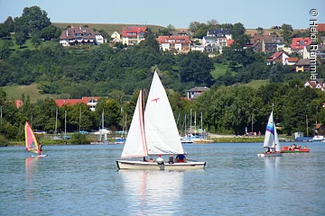 Ein weißes Segelschiff auf einem See. Daneben Windsurfer. Im Hintergund am Ufer Bäume. Etwas weiter hinten Wohnhäuser.