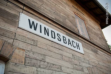 Nahaufnahme einer Hausfassade. An der Steinfassade hängt ein weißes Schild. Darauf in schwarzen Blockbuchstaben das Wort Windsbach.