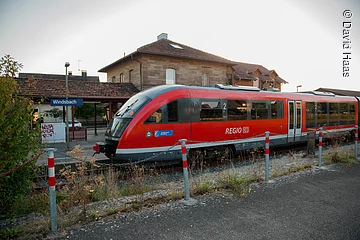 Ein kleiner Bahnhof. Auf dem einzigen Gleis steht ein roter Zug der deutschen Bahn. Dahinter ein Haus mit Steinfassade.