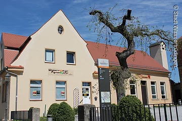 Ein Haus mit drei Ebenen und weißer Fassade. Davor ein Zaun. Über dem Eingang ein Schild mit der Aufschrift: Wassertrüdinger Figurentheater