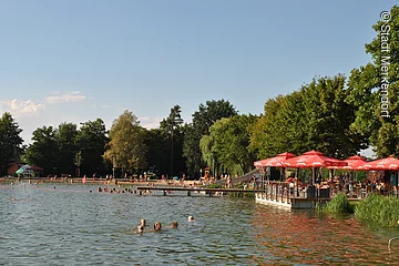 Ein Naturfreibad mit Steg. Im Wasser schwimmen Badegäste. Auf einer Holzterasse direkt am Wasser stehen rote Sonnenschirme.