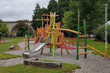 Ein Spieplatz mit großen Spielgeräten. Ein Klettergerüst mit Rutsche in bunten Farben. Im Hintergund grüne Bäume.
