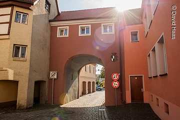 Ein Stadtor eingebunden zwischen zwei Häusern. Durch das Tor führt eine gepflasterte Straße der Altstadt. Über dem Torbogen drei kleine Fenster, dann folgt das Dach. Das Tor hat eine rötliche Fassade. Die Sonne spitzt über den Dächern hervor.