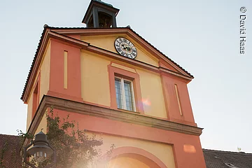 Ein Stadttor eingebunden zwischen einer Stadtmauer. Das Tor hat eine gelbe und rötliche Fassade. Ganz pben am Dachgiebel ist eine Uhr montiert. Durch das Tor führt eine gepflasterte Straße, außerdem scheint die untergehende Sonne durch das Tor.