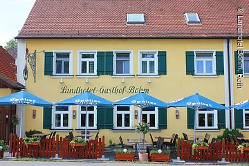 Teilansicht eines zweistöckigen Hauses im fränkischen Baustil mit gelber Fassade und grünen Fensterläden. Davor ein kleiner Biergarten mit braunem Zaun und blauen Sonnenschirmen.