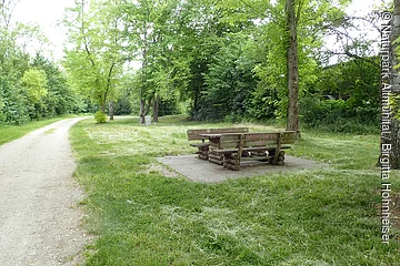 Unter dem Bäumen steht eine gemütliche Sitzgelegenheit aus Holz. Links führt ein geschotterter Weg entlang.