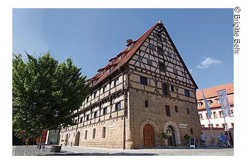 Ein großes Fachwerkhaus mit 6 Ebenen. Die Fassade der unteren zwei Stöcke besteht aus Stein, darüber braunes Fachwerk mit weißer Fassade.