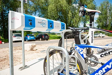 Eine Ladestation für Elektro-Fahrräder. Ein blau-weißer Ladebalken. Davor stehen zwei Rennräder.
