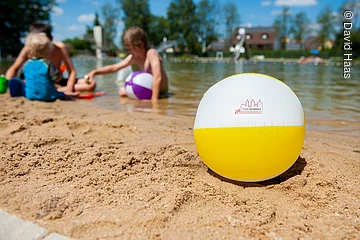 Im Vordergrund ein Wasserball am Strand. Dahinter spielen Kinder am Strandufer.