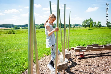 Ein junges Mädchen balanciert über einen Balken in Schlangenform. Dabei hält sie sich an Metallstangen fest. Im Hintergund grüne Wiesen.