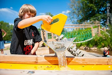 Ein Junge auf einem Wasserspielplatz. Er spielt mit einem Wasserspielplatz. Ein gelbes Gefäß aus welchem Wasser tröpfelt.