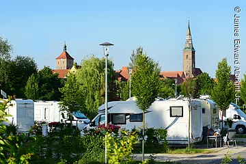 Auf einm Wohnmobilstellplatz stehen zwischen einzelnen Bäumen und Sträuchern weiße Wohmobile. Im Hintergrund versteckt erkennt man Häuser und einen Kirchturm die zwischen Bäumen hervorragen.