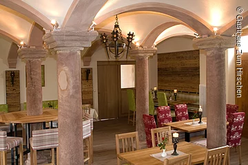 Festsaal des Landgasthauses Zum Hirschen mit eindruckvoller Gewölbedecke und im Raum verteilten Säulen. Im Raum stehen Holztische mit Stühlen. An der Decke hängt ein großer Kronleuchter.