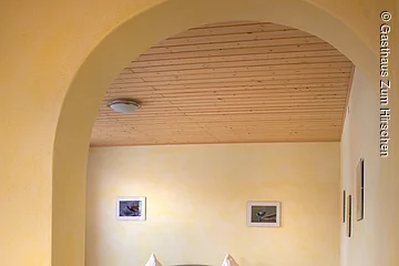 Durch einen Torbogen in der Wand fällt der Blick auf ein Doppelbett aus Holz mit blumiger Bettwäsche. Ein hellgelber Raum mit Dekce und Boden aus Holz.