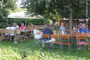 Ein Biergarten im Grünen. Tische und STühle aus Holz sthen unter grünen Bäumen auf einer Wiese. Der Garten ist umgeben von einer großen Hecke. Viele Gäste sitzen im Biergarten.
