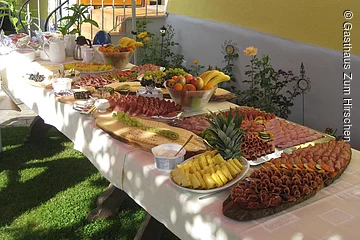 Großes Buffet mit verschiednen Leckereien. Ein langer Tisch mit weißer Tischdecke steht in einem Garten an einer gelben Hauswand. Auf dem Tisch viele vershciedne Platten mit Obst, Wurst oder Käse.