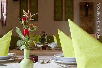 Nahaufnahme eines grün geschmückten Tisches. Auf der wei0en Tischdecke stehen Teller und Besteck sowie eine grüne Vase mit roter Rose zur Dekoration.