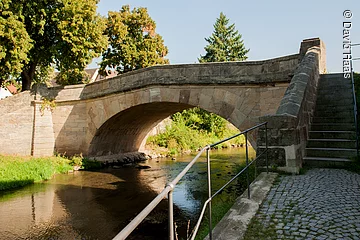 Eine Steinbrücke führt über einen Fluss. Im Hintergrund Bäume.