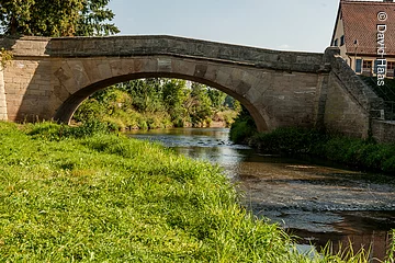 Eine Steinbrücke führt über einen Fluss. HInter der Brücke wachsen am Ufer Bäume. Am Ufer vor der Brücke eine grüne Wiese.
