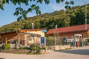 Das Gebäude aus Holz ist l-förmig gebaut und schön mit Pflanzen und Steinen angepflanzt. Hinter dem Campingplatz sieht man den Wald. Der Himmel strahlt himmelblau.