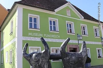 Außenansicht des Archäologischen Museums in Thalmässing. Grünes Haus mit schwarzem Dach. Davor steht eine Skulptur von menschlichen Figuren, die eine Platte tragen.