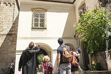 Eine Familie steht mit einem Mönch vor dem Eingang zum Kloster Plankstetten. Der Mönch erklärt der Familie etwas von der Geschichte des Gebäudes. Er hat seinen Arm gehoben und zeigt auf etwas.